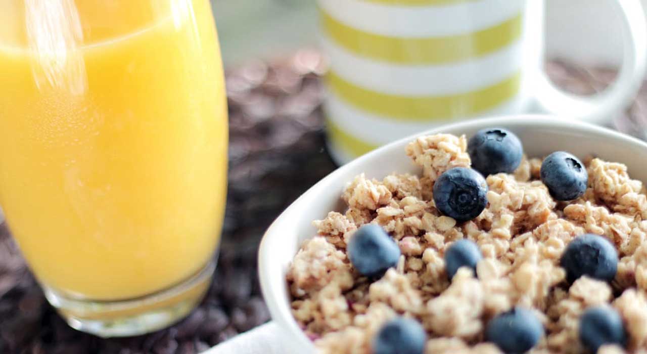 glass of orange juice next to a cup of tea and bowl of granola and blueberries