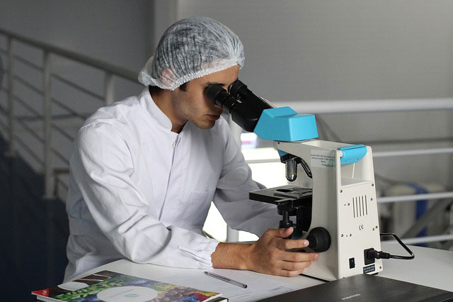 man dressed dressed in white with a hairnet looking into a microscope in a science lab