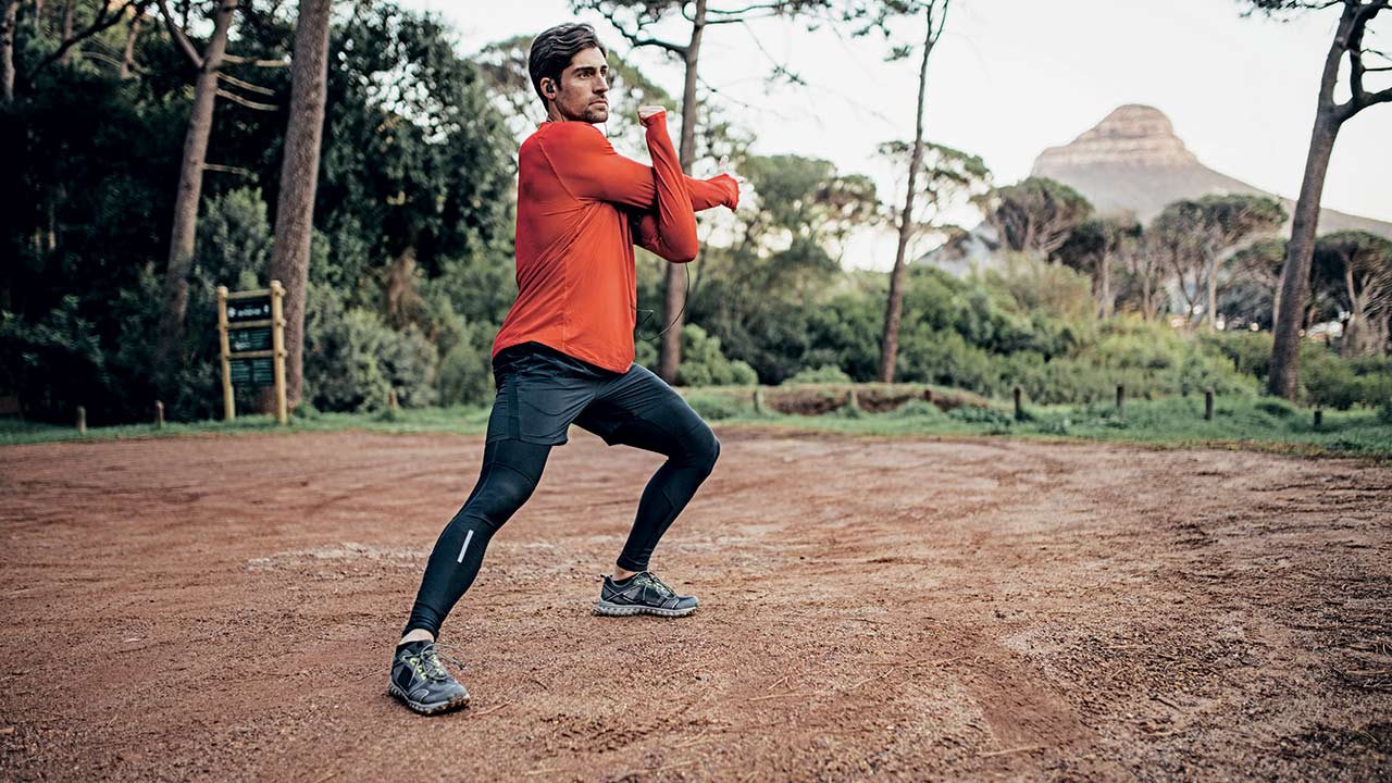 man in red tshirt with black thermal leggings stretching on a dirt track next to trees and bushes