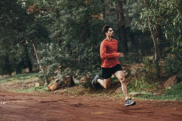 fit man in red tshirt and black shorts running on a dirt track next to some woods