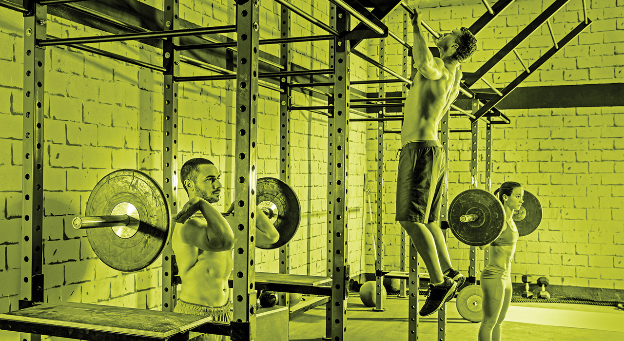 two muscular men in a gym one holding a barbell and the other performing a pull up