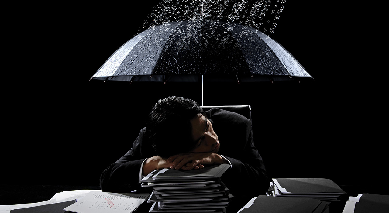a man sitting on a chair with his head on some books with an umbrella protecting him from the rain