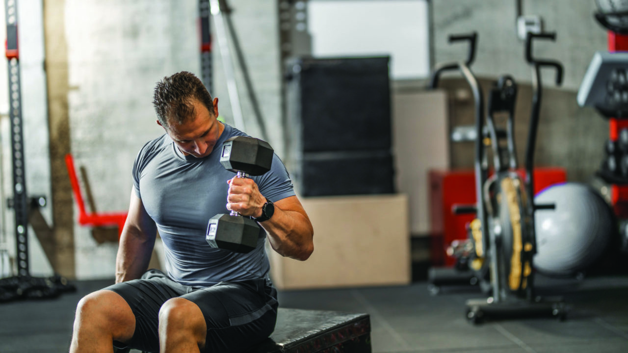 Muscular guy in sportswear lifting dumbbell while sitting on bench at cross training gym. Mature athlete using dumbbell during a workout. Strong man under physical exertion pumping up bicep muscle with heavy weight.